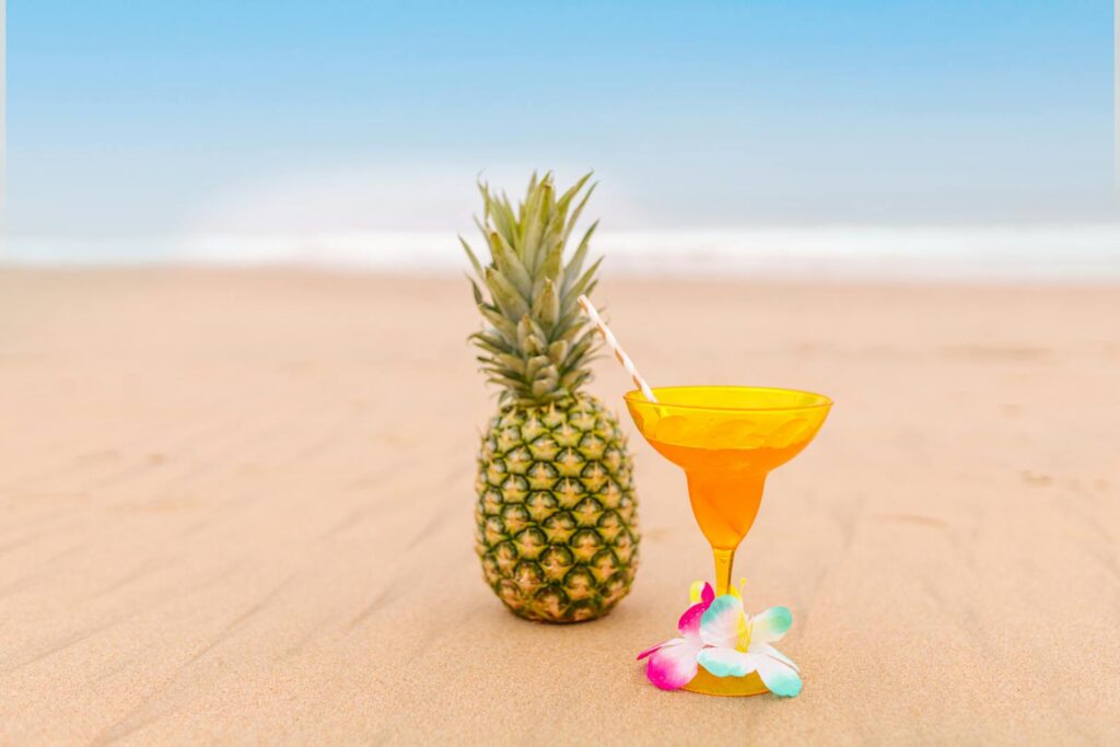 A vibrant pineapple cocktail with a straw on a sandy beach, evoking summer relaxation.